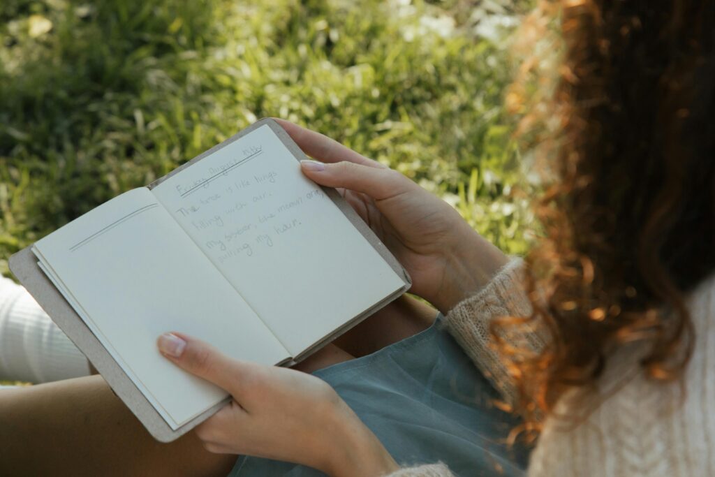 Adult with wavy hair reads a journal outdoors in a sunny park setting, reflecting on life.